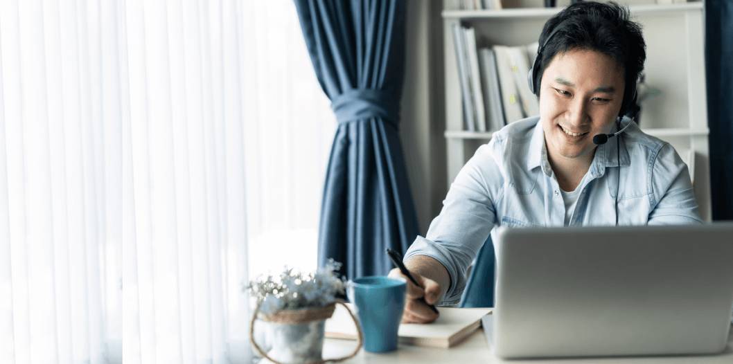 Man wearing headset smiling at a laptop screen, holding a pen to a notepad